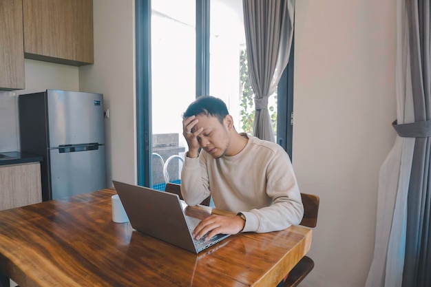 A young Asian man spends his time at home sitting in the dining room working on his laptop feel frustrated