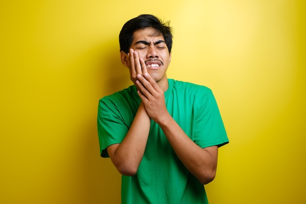 Young asian man touching his jaw having bad pain, toothache concept against yellow background