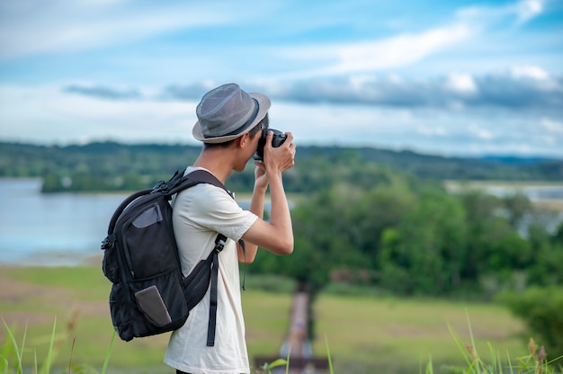 Young Asian man traveler wearing knitted hat with backpack taking a photo and looking at amazing lake. 