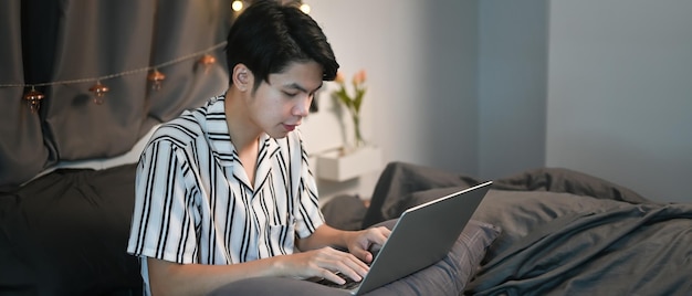 Young asian man using laptop while sitting on his bed at night