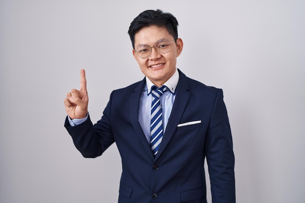 Young asian man wearing business suit and tie showing and pointing up with finger number one while smiling confident and happy.