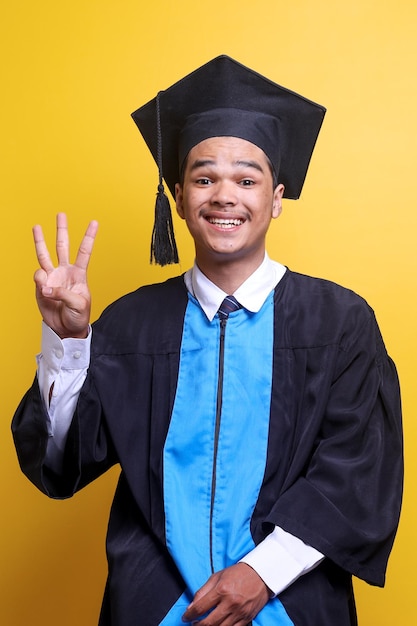 Young Asian man wearing graduation cap and ceremony robe with happy face showing number three and lo