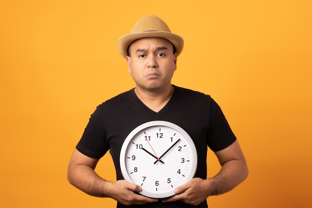 Young asian man wearing hat black shirt holding Big clock standing on isolated yellow background.He is face boring.