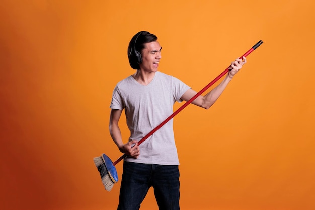 Young asian man in wireless, headphones playing rock song on mop as virtual guitar. Musician practicing, teenager using imaginable musical instrument, fun pastime, leisure activity