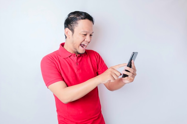 A young Asian man with a happy successful expression wearing red shirt and holding smartphone isolated by white background