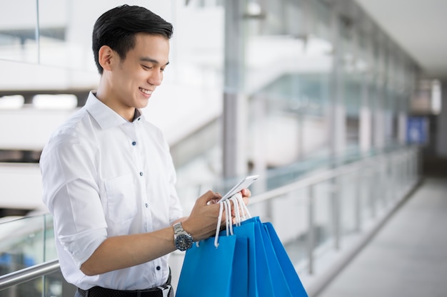 Young Asian man with shopping bags is using a mobile phone and smiling while doing shopping 