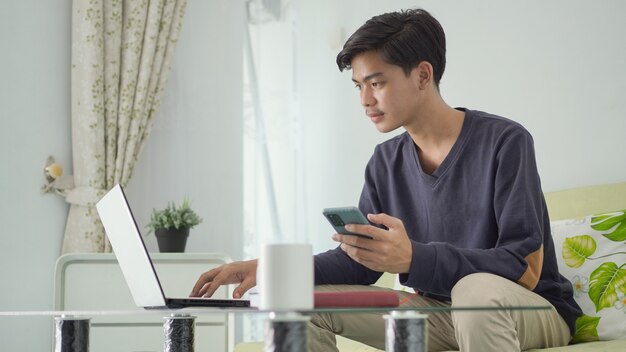 Young asian man working on his laptop while holding a cell phone at home