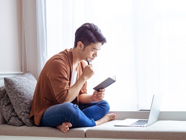 Young asian man writing in notebook and using laptop.