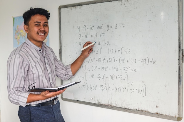 Young Asian math teacher holding books and explaining math formulas written on whiteboard