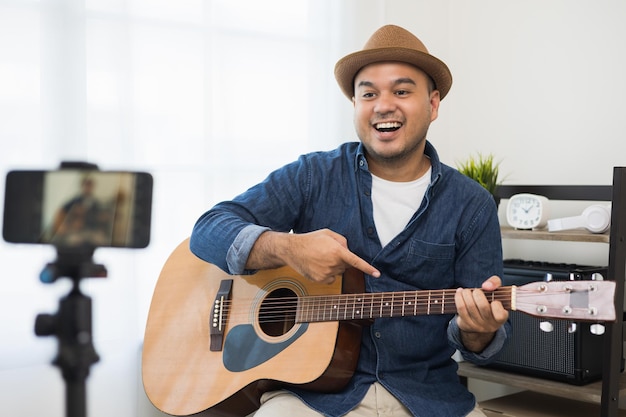 Young asian musician teaching the guitar class online live streaming with smartphone with student. Indian man playing acoustic guitar while singing a song and record by cell phone shaed online social