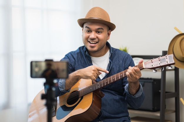 Young asian musician teaching the guitar class online live streaming with smartphone with student. Indian man playing acoustic guitar while singing a song and record by cell phone shaed online social