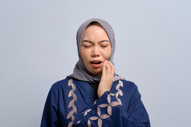 Young Asian Muslim woman feeling pain holding her cheek with hand suffering from severe toothache isolated over white background