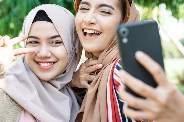 Young asian muslim woman in head scarf meet friends and using phone in the park for selfie or video calling