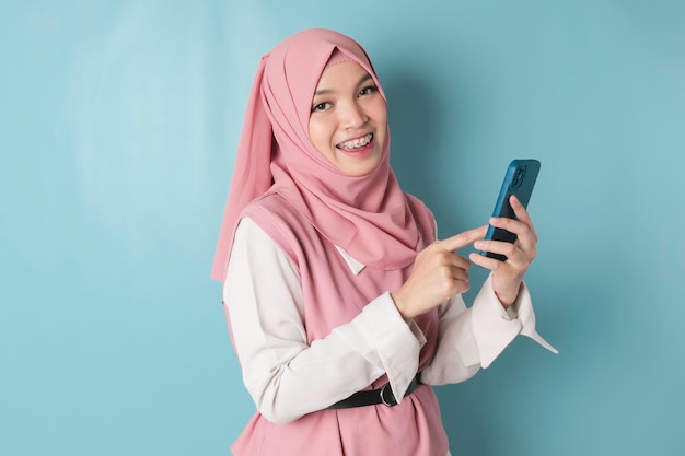 A young Asian Muslim woman wearing pink hijab is smiling while holding her smartphone