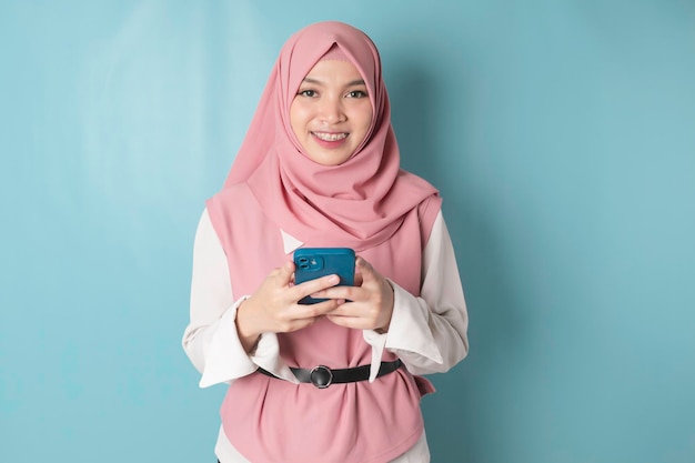 A young Asian Muslim woman wearing pink hijab is smiling while holding her smartphone