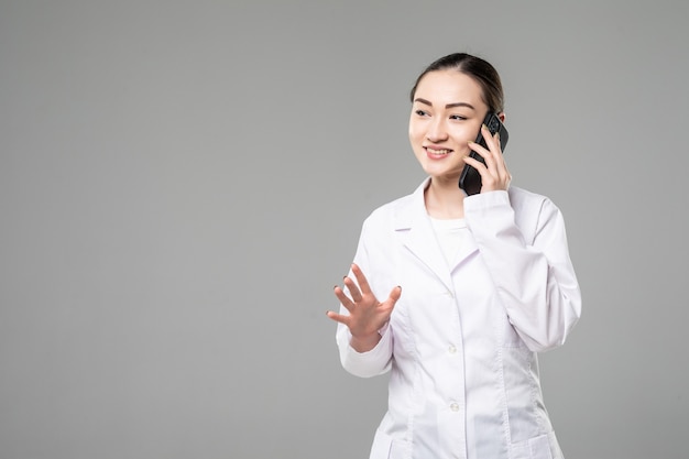 Young asian nurse with a stethoscope talking on the phone, isolated over white wall.