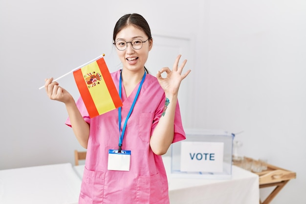 Young asian nurse woman at political campaign election holding spain flag doing ok sign with fingers smiling friendly gesturing excellent symbol