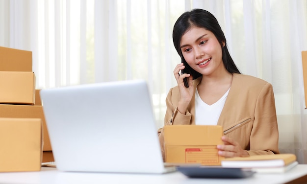 Young asian smart and happy woman in casual dress working from home with laptop computer and smart mobile phone for online purchase or shopping order with box packaging on table (new normal concept)