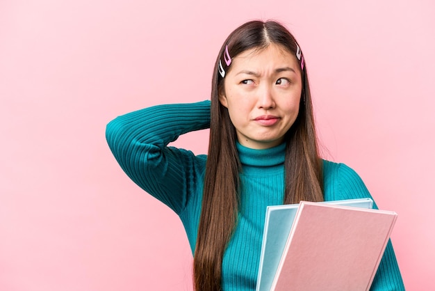 Young asian student woman holding books isolated on pink background touching back of head thinking and making a choice