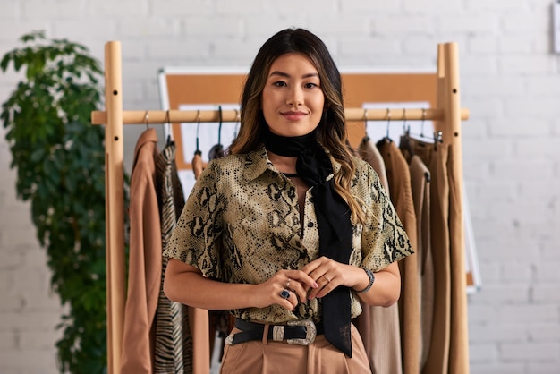 young asian stylist in trendy casual attire smiling at camera near rack with clothes in atelier