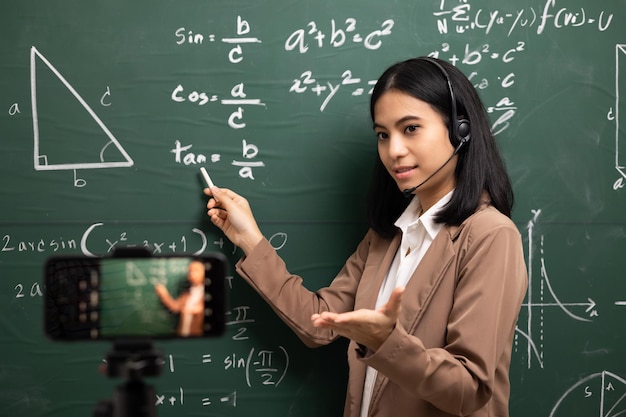 Young asian teacher woman standing looking at camera and video conference with student Female teacher training the mathematics in classroom from live stream with smartphone online course