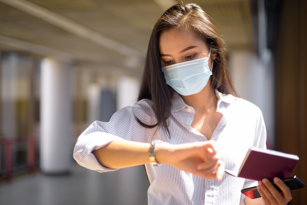 Young Asian tourist woman with mask checking time while holding passport at the airport