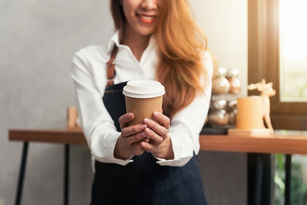 Young asian woman barista holding a disposable coffee cup with smiling face at cafe background