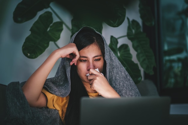 Young asian woman blowing nose and sneeze into tissue paper while working with laptop at night experiences allergy and flu symptoms health care concept