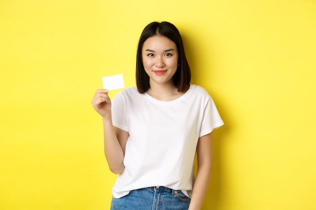 Young asian woman in casual white t-shirt showing plastic credit card and smiling at camera, yellow background.