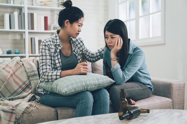 young asian woman comforting her depressed friend in living room at home while sitting on comfort couch sofa. two girls upset talking with bottles of beer and drinks on table. heartbreaking female.