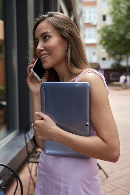 Young asian woman in dress standing outdoors with laptop calling by smartphone