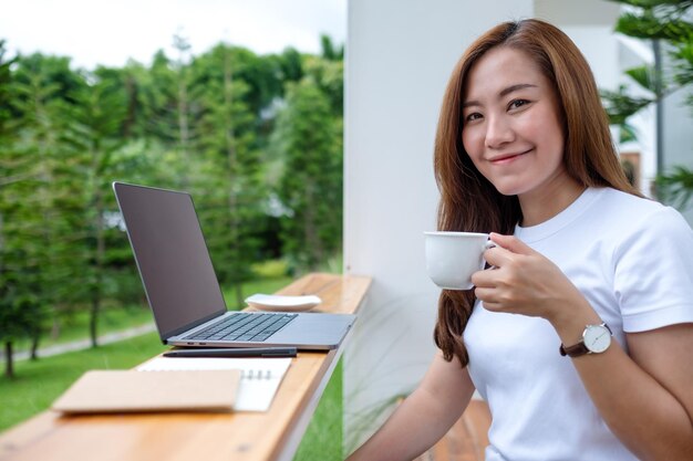 A young asian woman drinking coffee while using and working on laptop computer in the outdoors