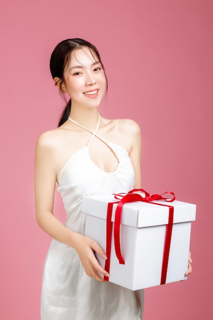 Young Asian woman gathered in ponytail with natural makeup on face and clean fresh skin wearing white camisole on isolated pink background Portrait of cute female model holding gift box in studio