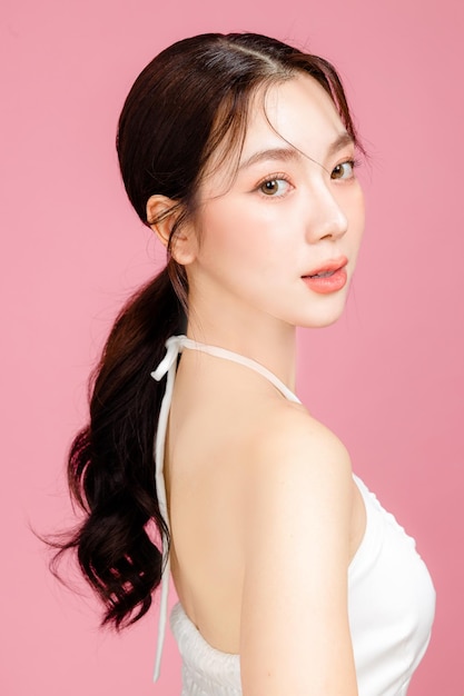 Young Asian woman gathered in ponytail with natural makeup on face have plump lips and clean fresh skin wearing white camisole on isolated pink background Portrait of cute female model in studio