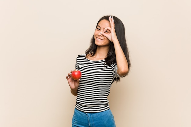 Young asian woman holding an apple excited keeping ok gesture on eye.