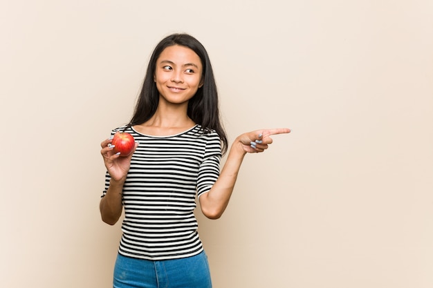 Young asian woman holding an apple smiling and pointing aside, showing something at blank space.