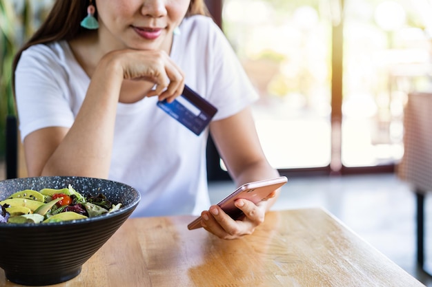 Young asian woman holding a credit card and using smart phone for make an online shopping payment at restaurant