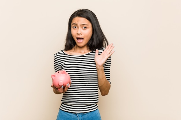 Young asian woman holding a piggy bank surprised and shocked.