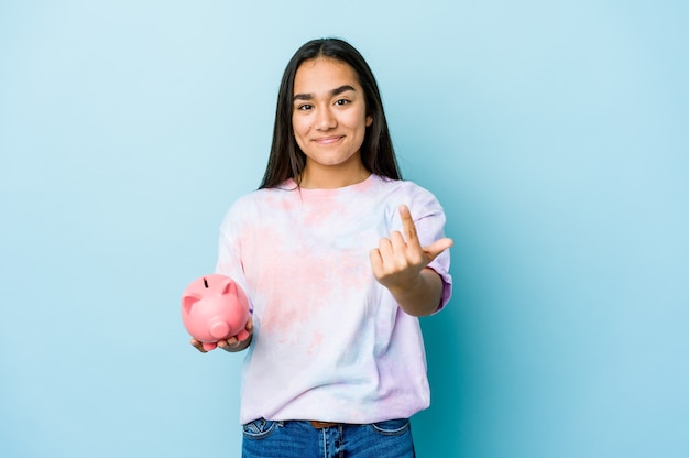 Young asian woman holding a pink bank over isolated wall pointing with finger at you as if inviting come closer.