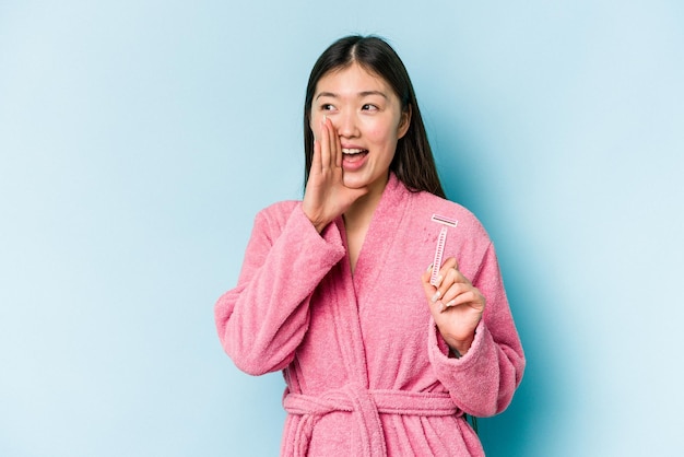 Young asian woman holding razor blade isolated on blue background shouting and holding palm near opened mouth