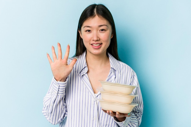 Young asian woman holding Tupperware isolated on blue background smiling cheerful showing number five with fingers