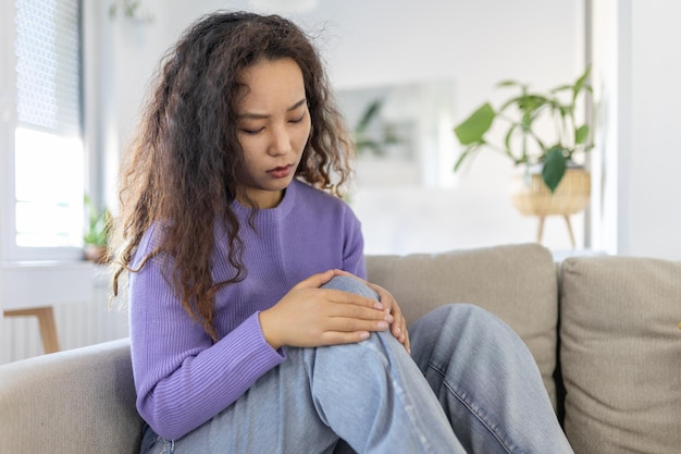 Young Asian woman is sitting on the couch at home has pain in the knee joint holding her leg osteoarthritis concept