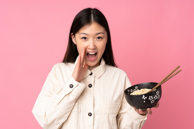 Young asian woman on pink wall shouting with mouth wide open while holding a bowl of noodles with chopsticks