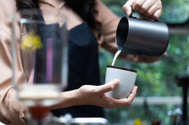 Young Asian woman pouring milk into coffee making late coffee. Professional barista preparing coffee on counter