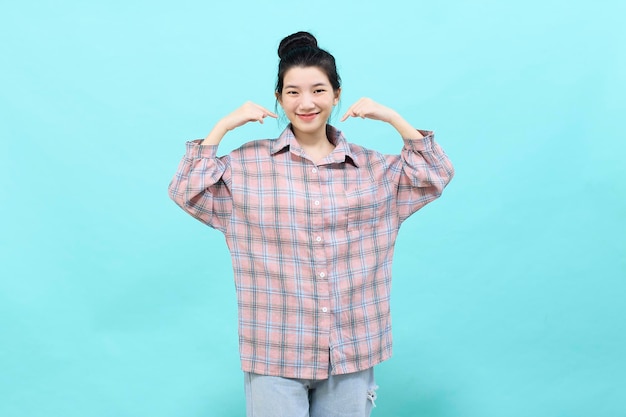 A young Asian woman pretty pointing looking at ads wearing a plaid shirt standing and feeling happy and confident on blue background