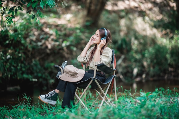 Young asian woman sit in a chair near the stream listening to music from tablet with wireless headphones happily while camping in the woods