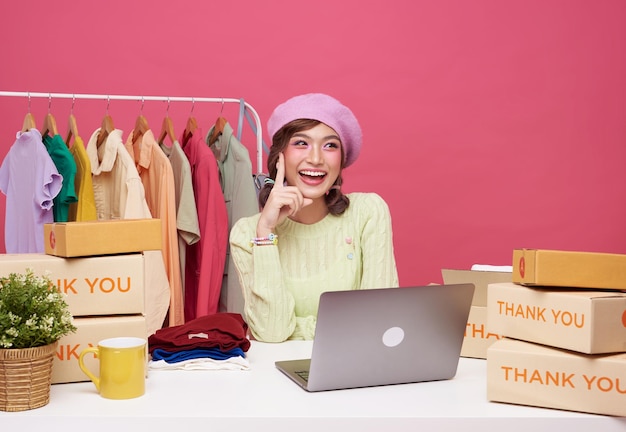 Young asian woman startup small business freelance sale fashion clothing with parcel box and computer laptop on table and sitting isolated on pink background Online marketing and delivery concept