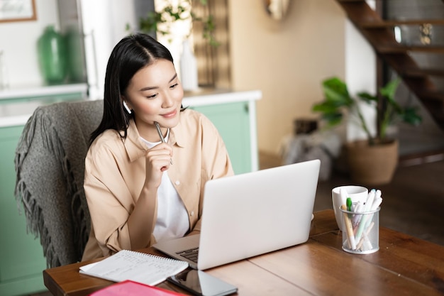 Young asian woman studying student listening webinar from home wearing earphones and looking at lapt...