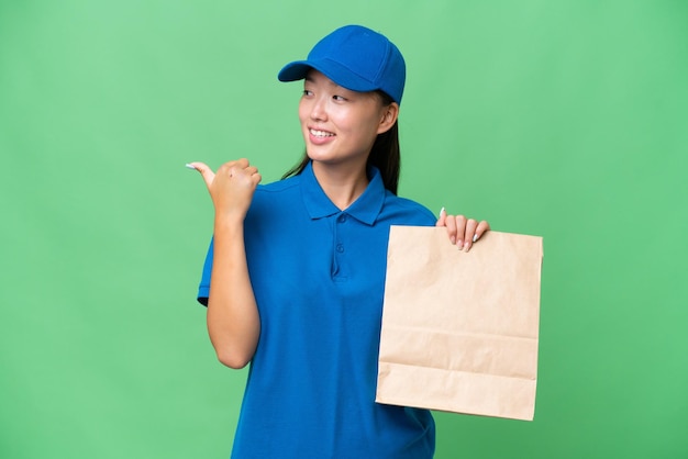 Young Asian woman taking a bag of takeaway food over isolated background pointing to the side to present a product