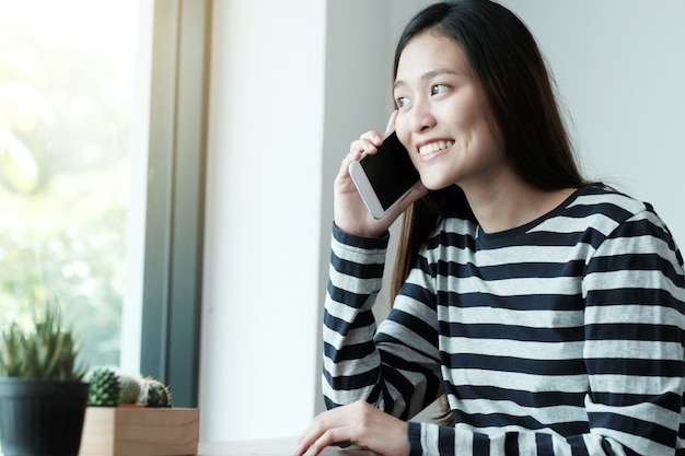 Young asian woman talking smart phone while sitting by window cafe background